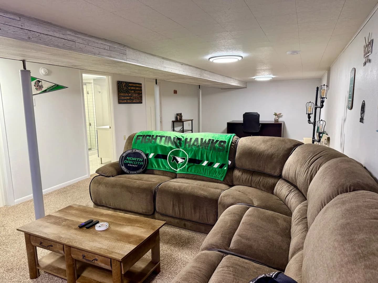 Brown sectional with a green Fighting Hawks blanket and hockey puck cushion, with a desk behind it