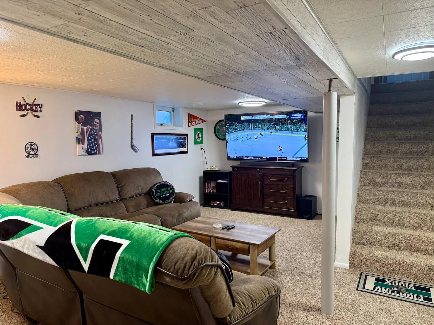 Basement sectional facing a large TV, with hockey photos, pennants, and a stick on the wall