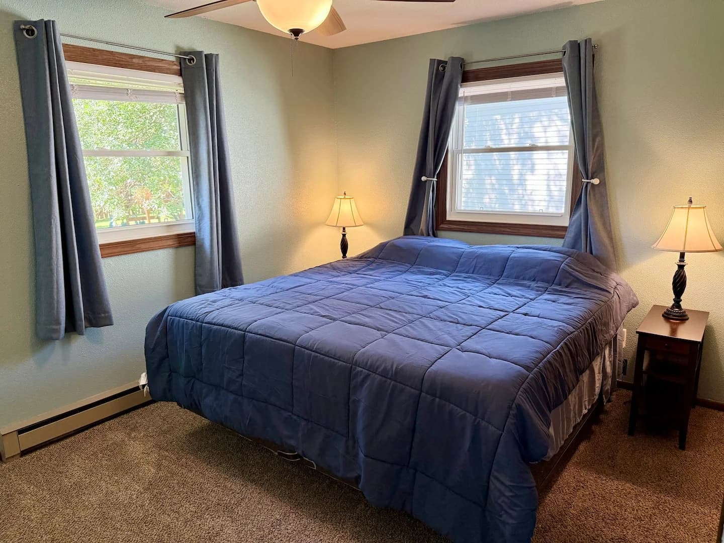 King bed with a blue quilt, two windows with dark curtains, and bedside lamps
